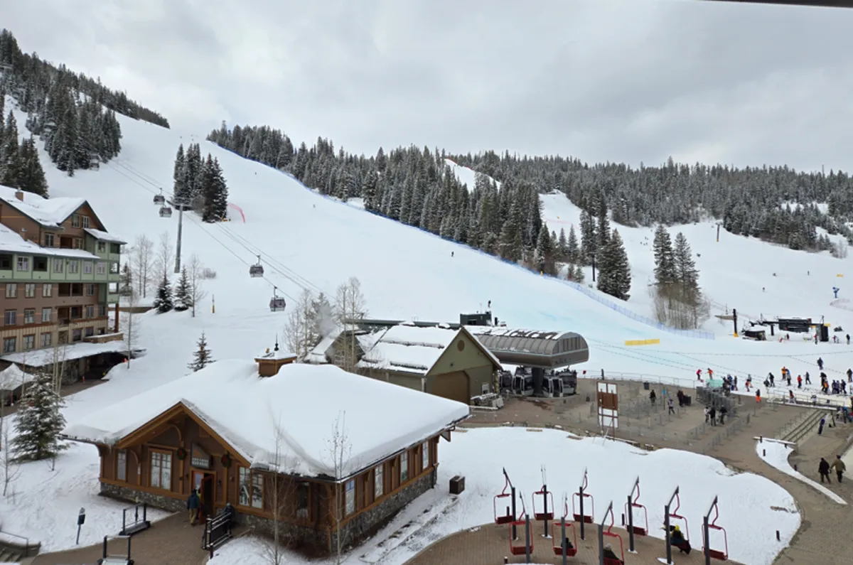 A vibrant winter scene showing ski slopes bustling with activity, with people skiing and snowboarding down the runs. The lodge buildings are visible at the base of the slopes, and ski lifts transport guests up the mountain. The backdrop features snow-covered mountains under a bright sky, evoking the excitement and energy of a ski resort in full swing.