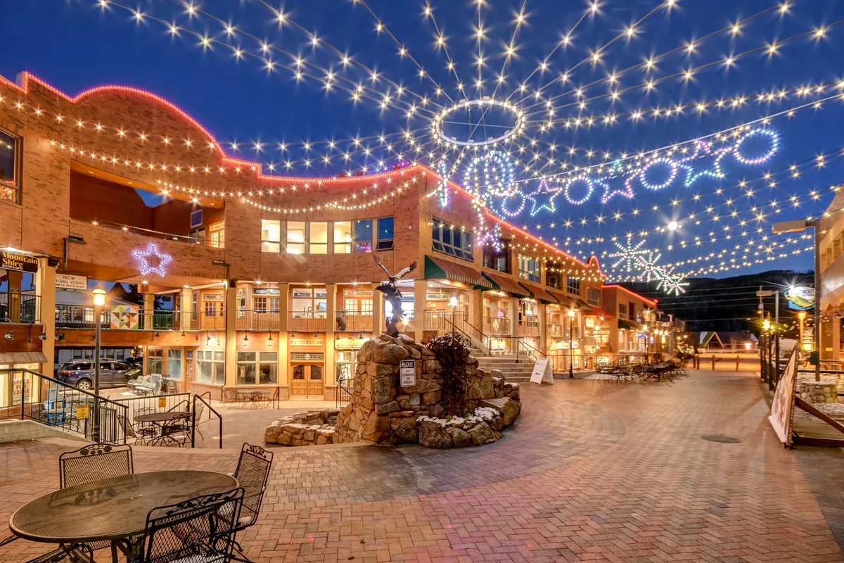 A charming evening scene of a central plaza with decorative holiday lights strung overhead, a central fountain surrounded by outdoor seating, and the facades of shops and restaurants framing the scene. People are seen dining outdoors, enjoying live music, and participating in a community event under the festive lights.