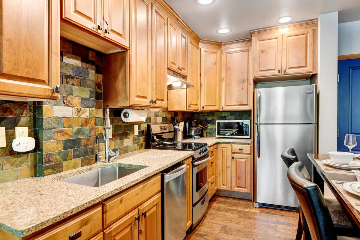 A warm, cozy kitchen featuring wooden cabinets, a granite countertop, a stone backsplash, and sleek stainless steel appliances. The scene suggests an inviting space for both meal prep and socializing, with plenty of room for multiple cooks or casual conversation at the island.