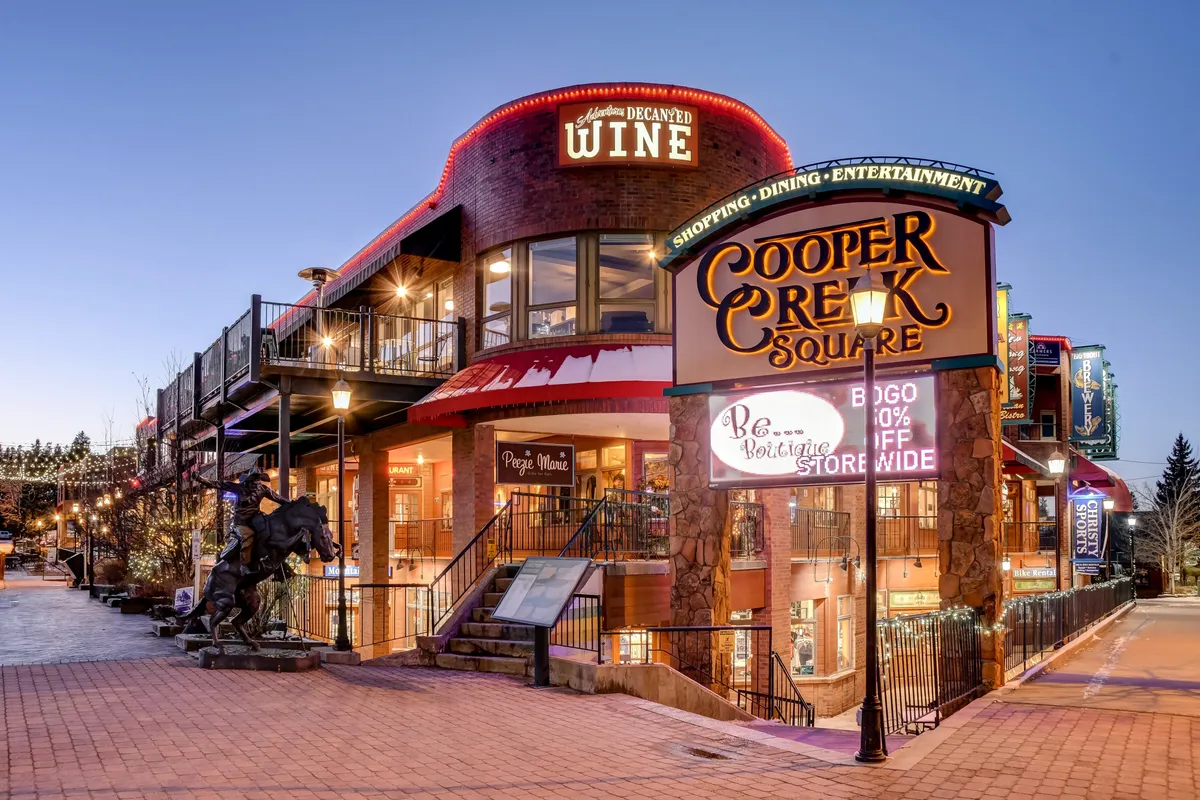 The exterior view of Cooper Creek Square, highlighted by its iconic sign, a welcoming wine shop entrance, and a staircase leading to additional shops and eateries. Pedestrians are visible, suggesting movement and activity, while banners and subtle festive decor hint at seasonal events and local nightlife.