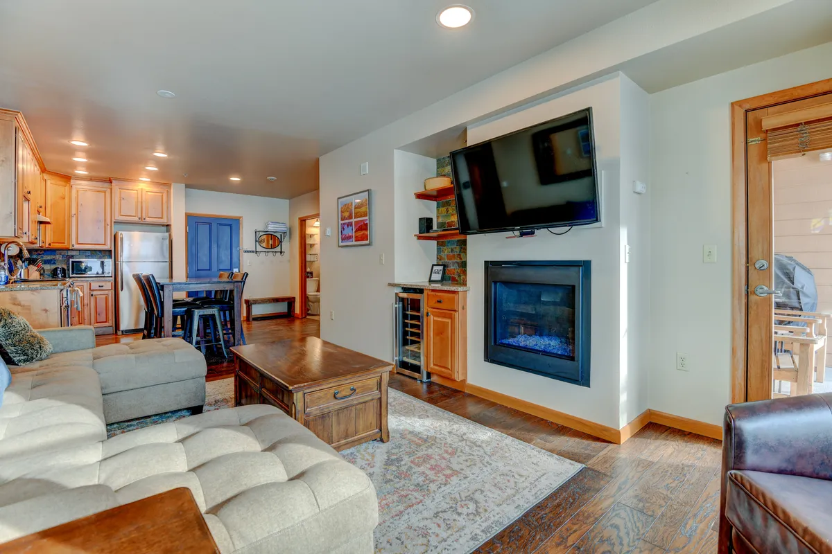 This inviting interior scene features a comfortable sectional sofa facing a stone fireplace, with a mounted television above and a wooden dining table nearby. Large windows let in natural light and offer a peek out to the deck or patio area, seamlessly blending indoor and outdoor living. The room is designed for relaxation and social gatherings, with quality furnishings and cozy accents.