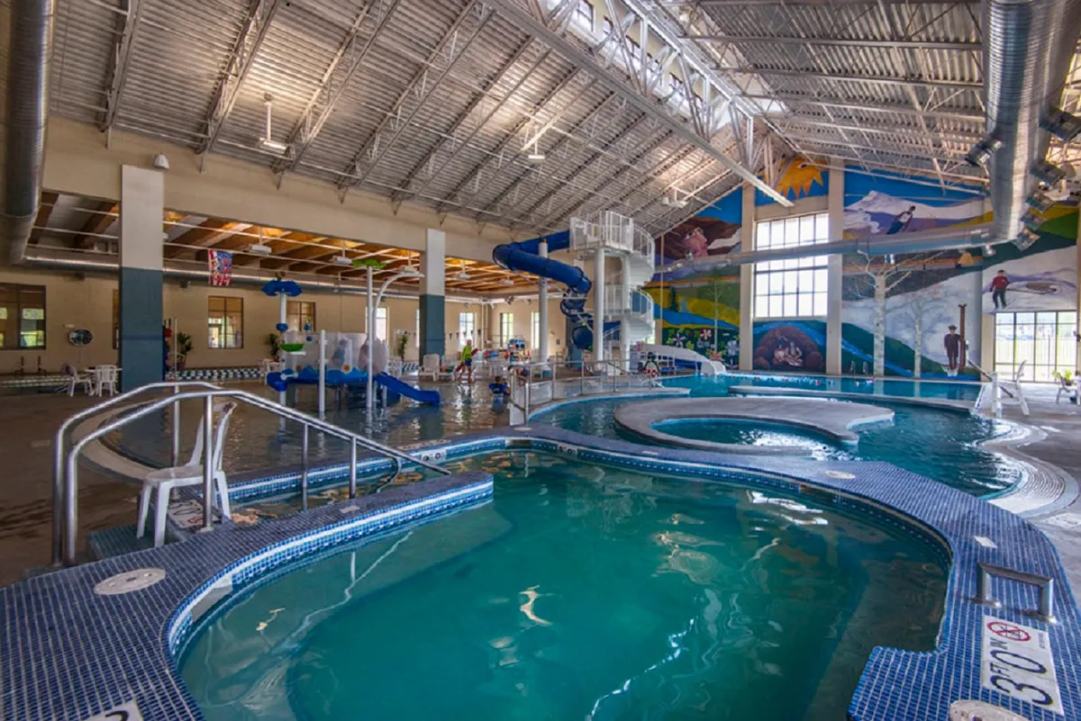 An energetic indoor pool scene with a large water slide, children splashing in the pool, colorful murals, and relaxed adults enjoying poolside seating. The joyful atmosphere hints at the resort's rec center amenities perfect for families on hot summer days.