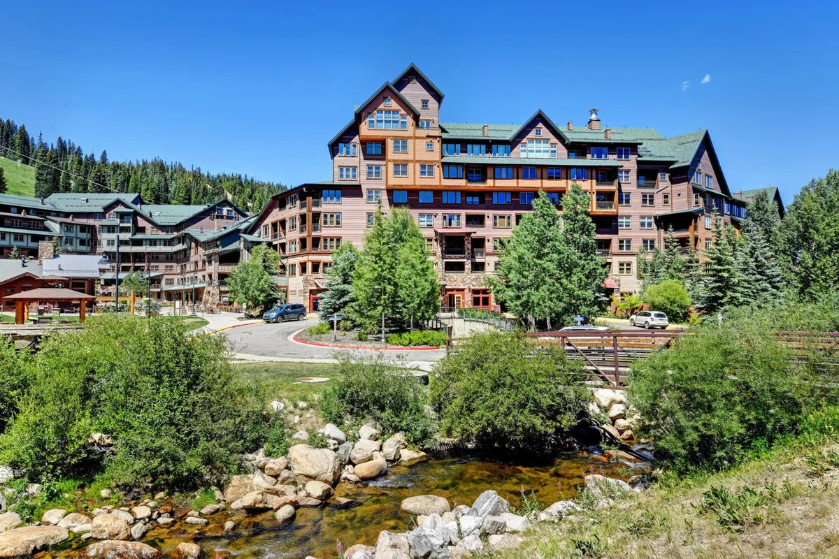 A relaxing exterior view of a large mountain lodge nestled among tall trees, with a gently flowing stream in the foreground and towering mountains in the distance. The scene includes people hiking and gathering outdoors, suggesting both exploration and relaxation in a natural setting.