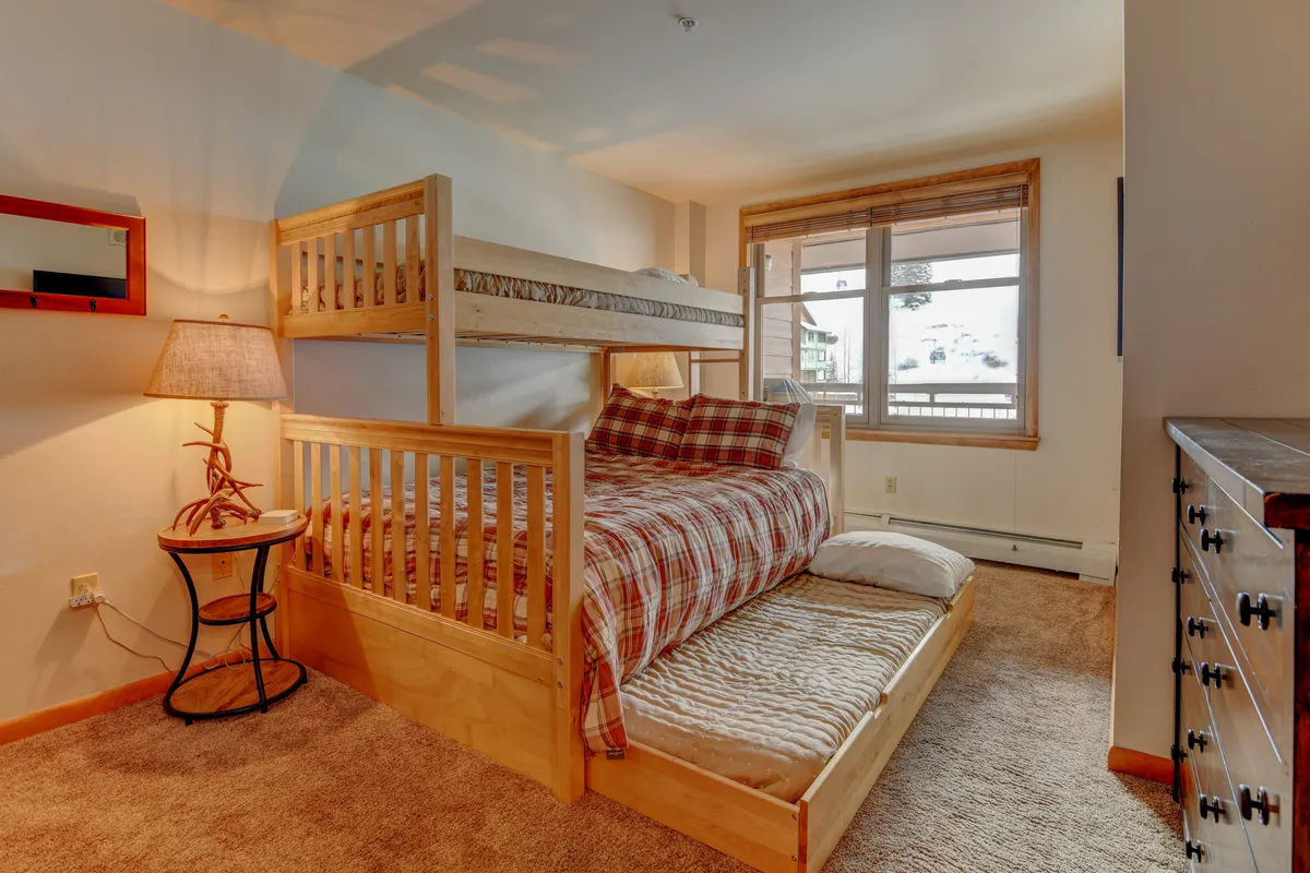 A family-friendly bedroom showcasing a twin-over-queen bunk bed with plaid bedding, a trundle, and a nightstand with a softly glowing lamp beside a window letting in natural light.