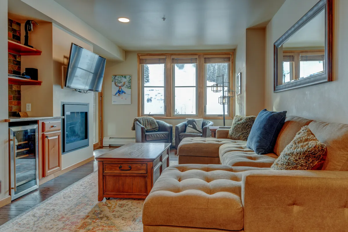 A beautifully furnished living room with plush, comfortable couches centered around a glowing fireplace, framed by expansive windows that showcase breathtaking snow-covered mountain views. The warm, inviting space features a stylish coffee table and plenty of room for relaxing or enjoying time with friends and family.