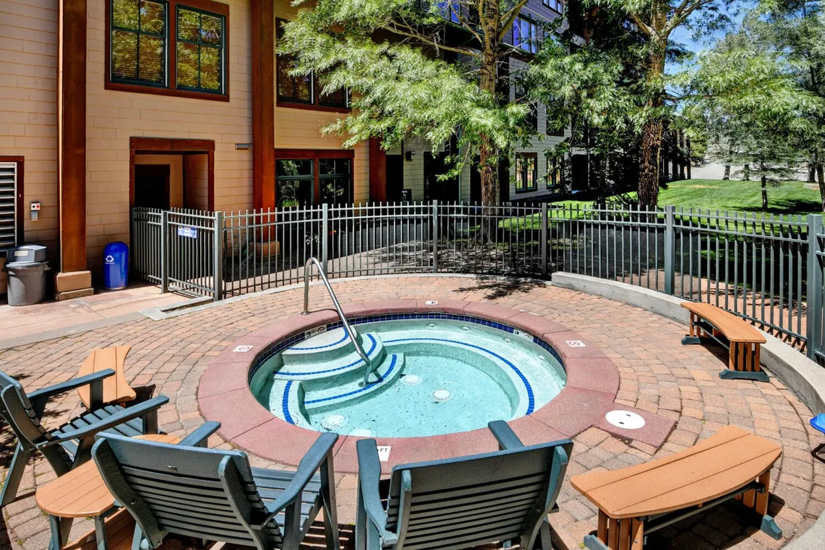 An inviting outdoor hot tub area surrounded by wooden benches and lush greenery. The setting exudes tranquility and warmth, perfect for unwinding after a day on the slopes. Steam rises from the hot tub, and the ambiance suggests a communal, relaxing retreat.