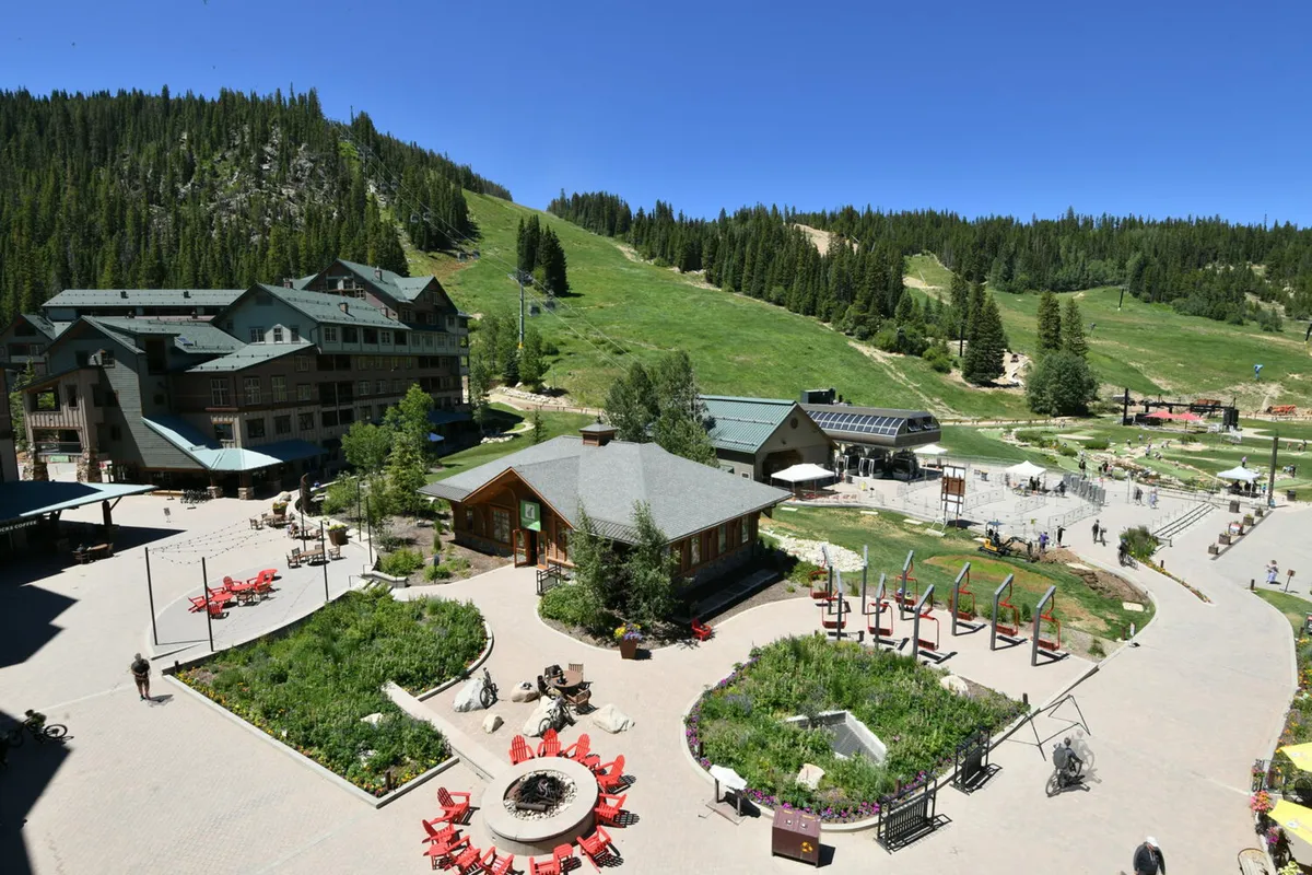 A cheerful exterior shot of the lodge with outdoor seating areas surrounded by lush greenery and mountains in the background. Groups of guests relax by the fire pit or enjoy outdoor dining, hinting at both adventure and relaxation within a beautiful mountain setting.
