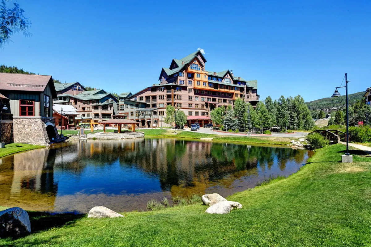 A tranquil scene featuring a large multi-story resort building beside a calm pond, lush green lawns, and mature trees. The outdoor spaces appear welcoming for family picnics, strolls, or relaxation between adventures.