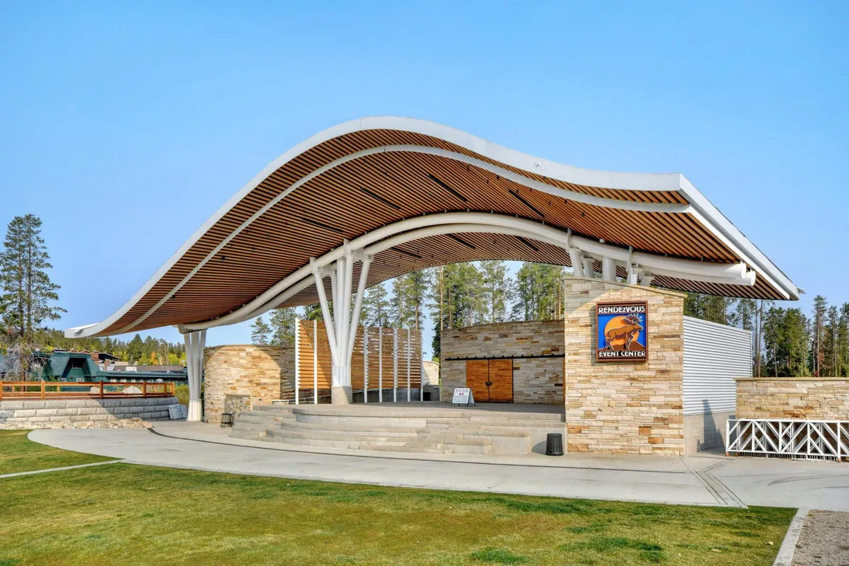 The exterior of the Hideaway Park amphitheater, with its curved roof, stone facade, and spacious outdoor stage area. The scene suggests a lively atmosphere for community events, concerts, and family gatherings against a mountain backdrop.