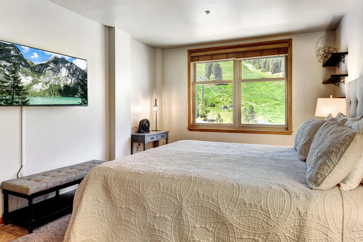 A serene bedroom featuring a queen-sized bed dressed with decorative bedding, a large window framing a breathtaking mountain view, and a nightstand with a lamp. The television displays a scenic landscape, enhancing the room's tranquil atmosphere.