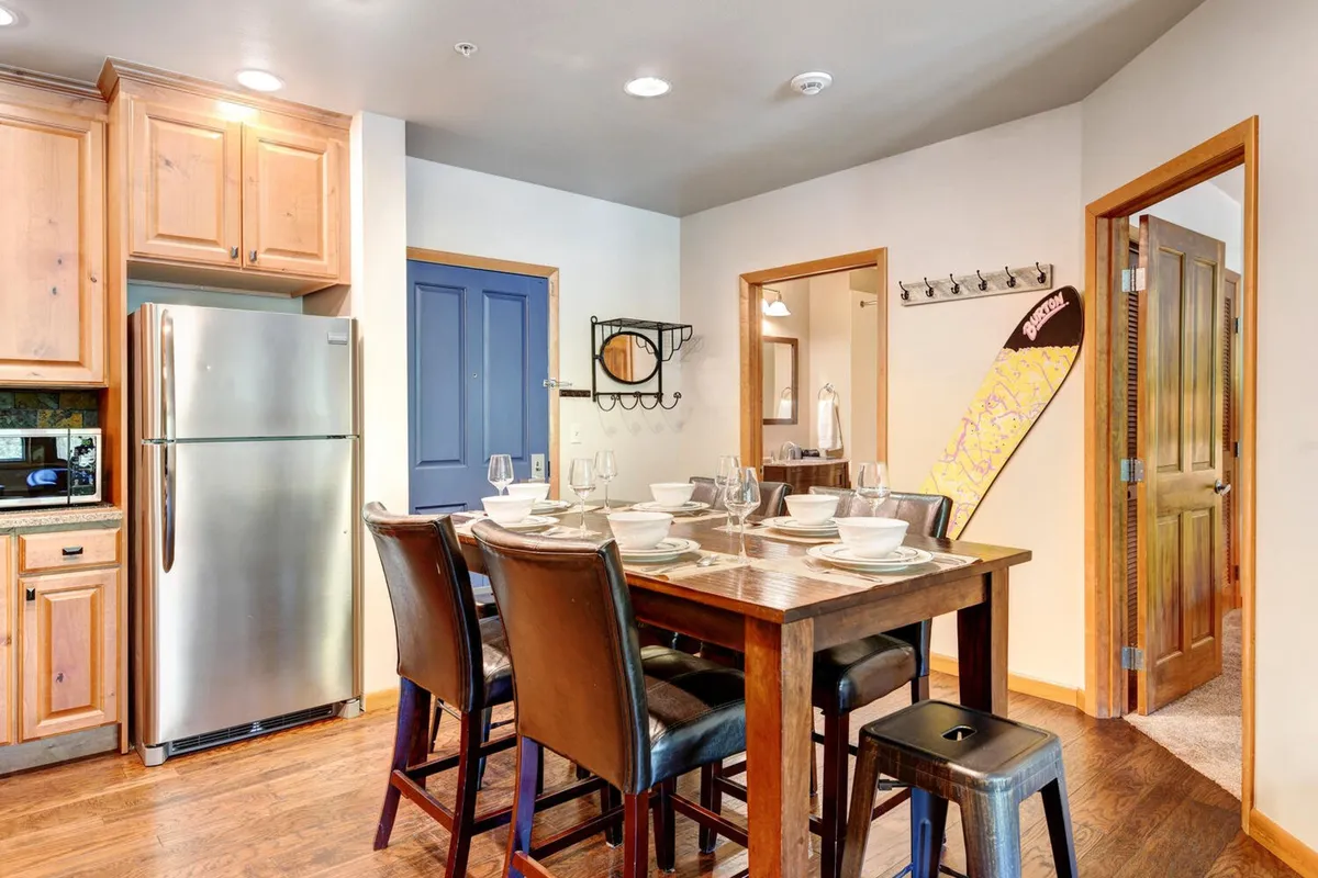 A dining area adjacent to the kitchen, with a spacious table set for a family gathering. Visible are kitchen appliances and a blue door, with a cozy, inviting mood that hints at memorable meals, casual gatherings, and board game fun with loved ones.