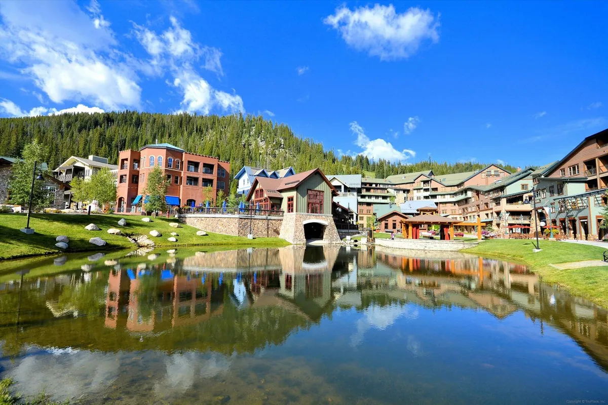 A vibrant exterior scene featuring a pond with a picturesque bridge, surrounding buildings, and a mountain backdrop. Guests can be seen strolling along the water, dining outdoors, or relaxing by the pond, suggesting a variety of seasonal activities and community events.