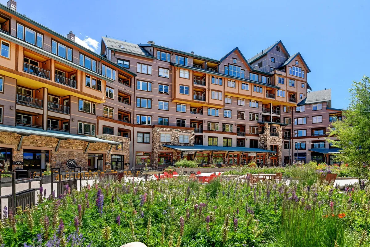 A lively exterior scene featuring a vibrant flower garden wrapping around a modern building with an inviting outdoor seating area. Several groups are dining al fresco, while others relax and engage in conversation amid the garden setting. The sunset creates a warm glow, and balconies overlook the scene, suggesting a bustling community atmosphere.