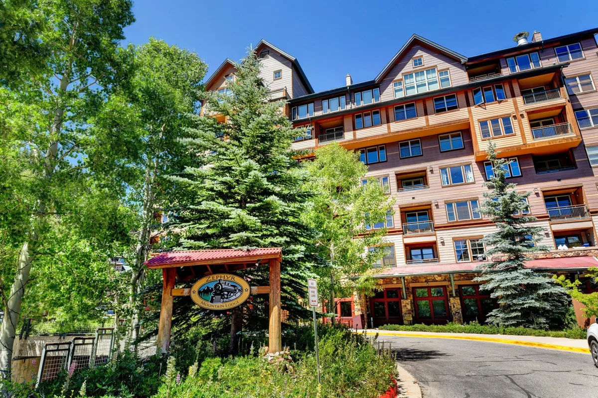This exterior shot features the inviting facade of Zephyr Mountain Lodge, framed by surrounding trees and a landscaped pathway. The building's architecture and signage are clearly visible, establishing a strong sense of place and arrival.